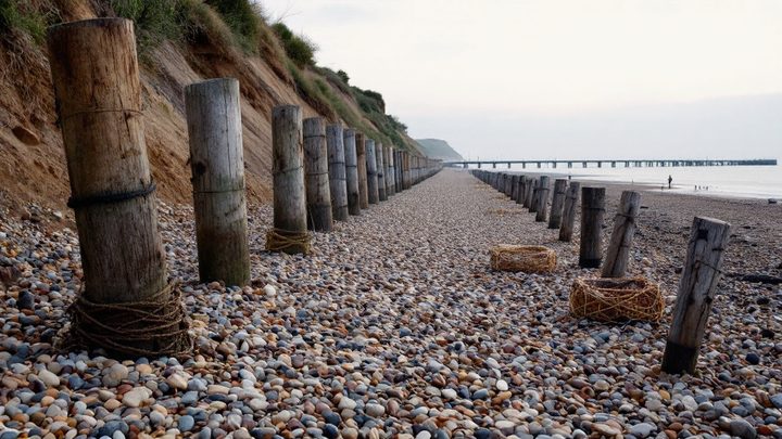 A shingle beach with wooden groynes and lobster pots, with a pier in the distance